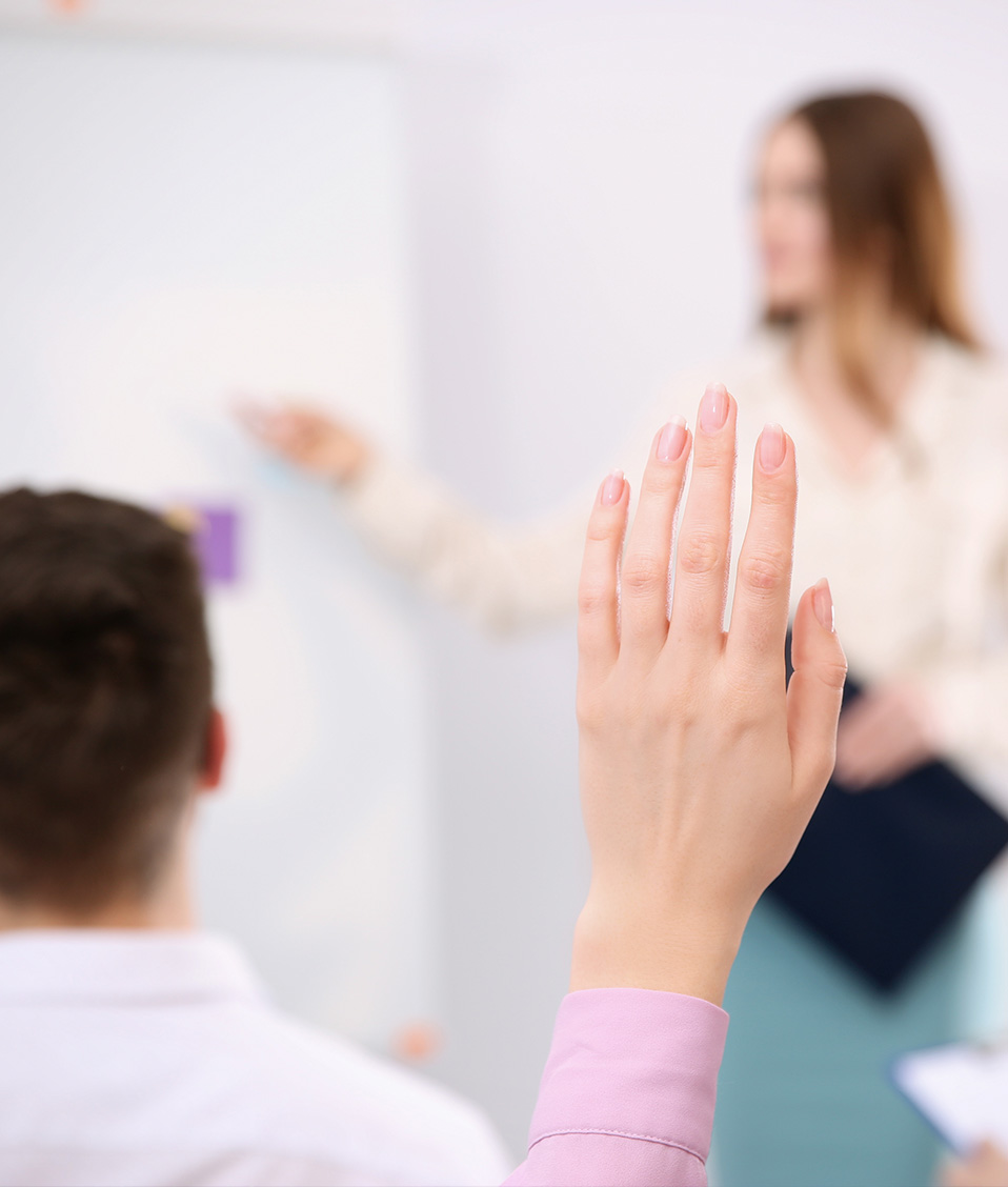 Mujer exponiendo al fondo en una reunión con varias personas que levantan la mano para hacer una pregunta