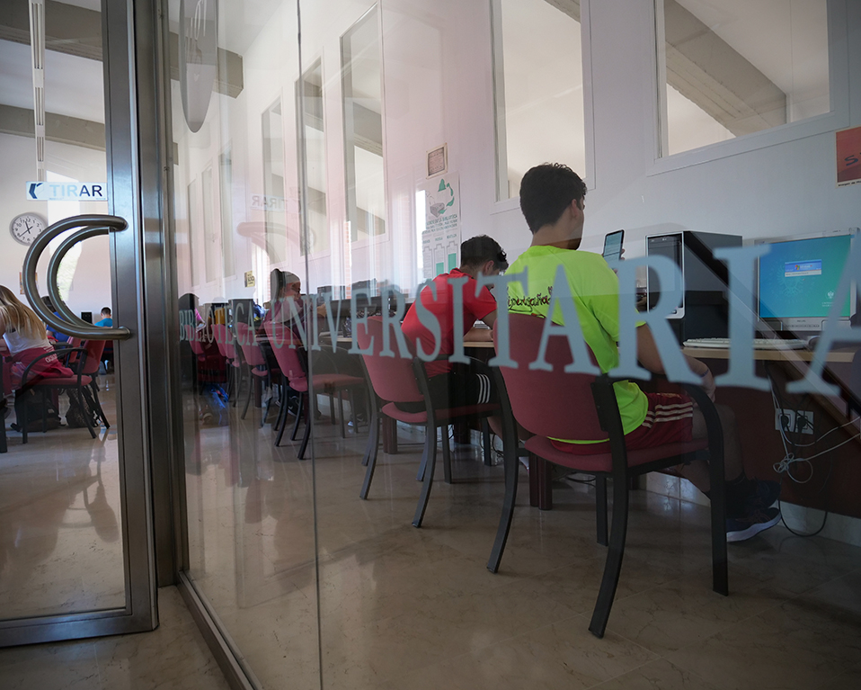 Puerta de cristal de entrada a la biblioteca de la Facultad de Ciencias del Deporte con estudiantes en ordenadores de libre acceso