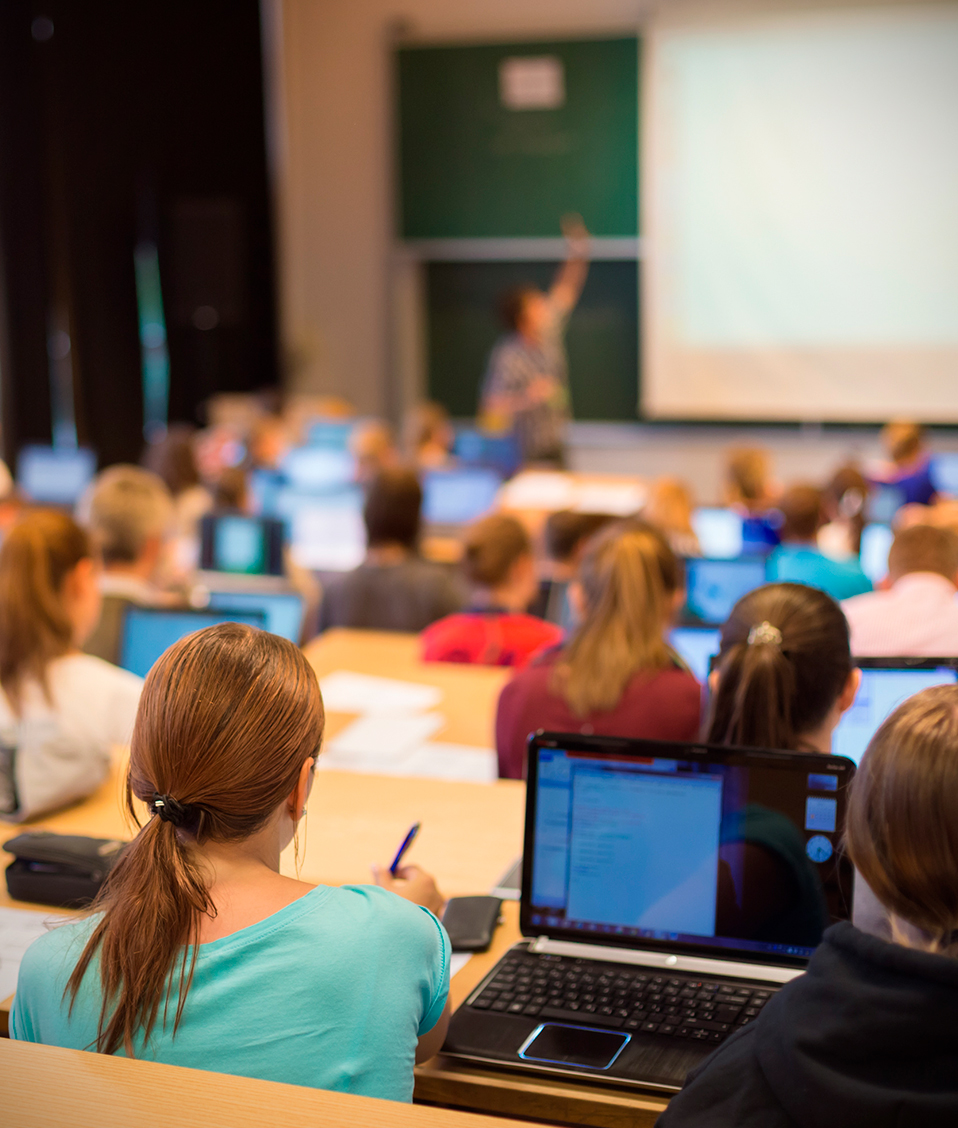 La imagen muestra una clase magistral en la que los estudiantes que ocupan los diversos asientos, hacen uso de sus ordenadores personales para seguir la explicación