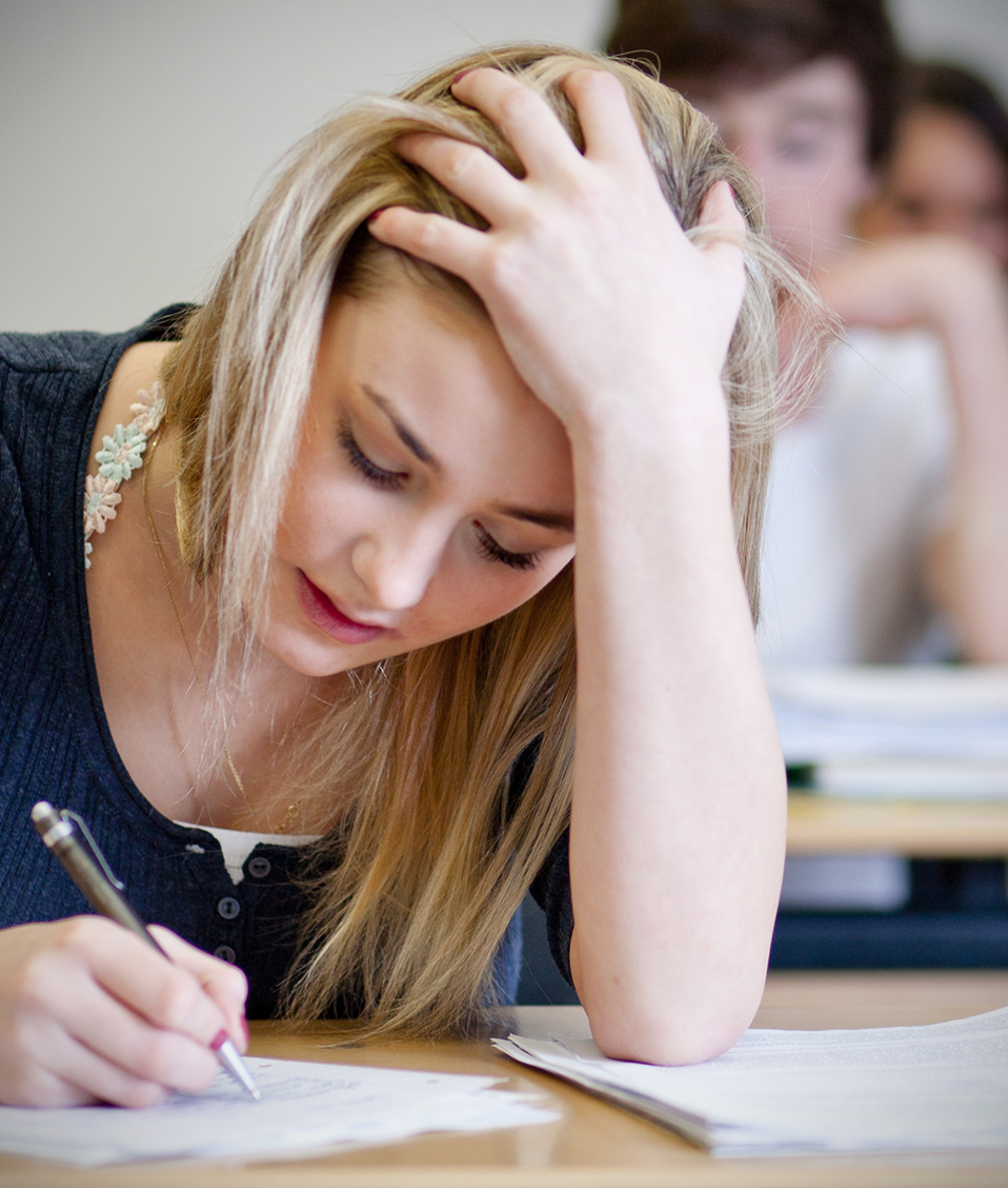 Una estudiante toma apuntes sentada en un pupitre, a sus espaldas, el resto de sus compañeros se encuentran realizando la misma tarea