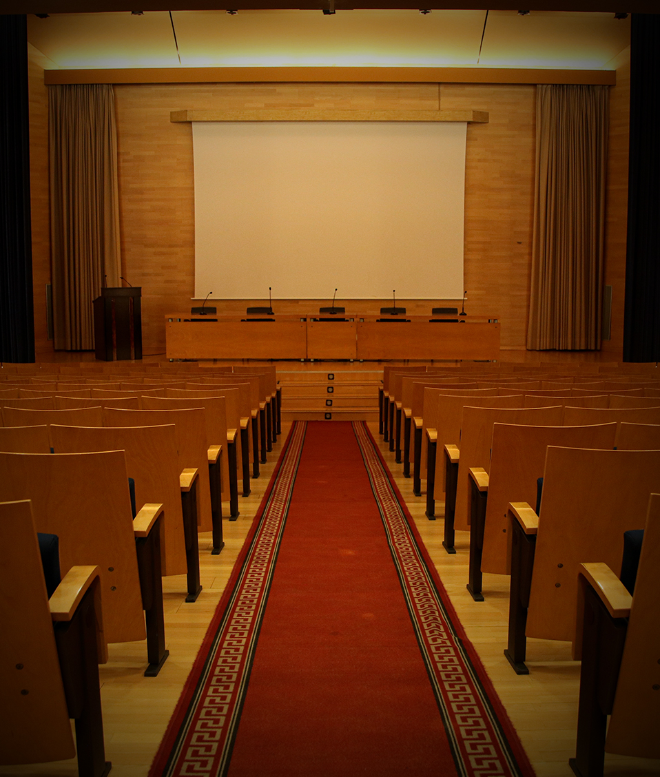 Salón de actos con un pasillo en medio, butacas a ambos lados y escenario con una mesa al fondo en donde hay micrófonos