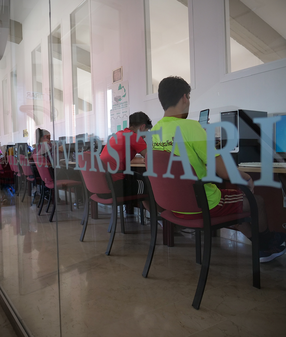 Puerta de cristal de entrada a la biblioteca de la Facultad de Ciencias del Deporte con estudiantes en ordenadores de libre acceso