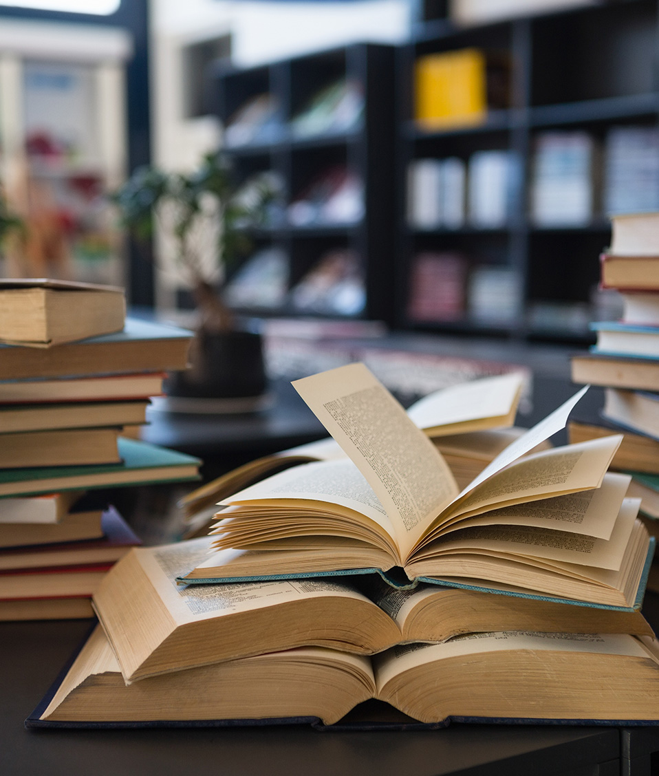 Biblioteca con una mesa en primer plano con varias torres de libros apilados, la del centro con los libros abiertos