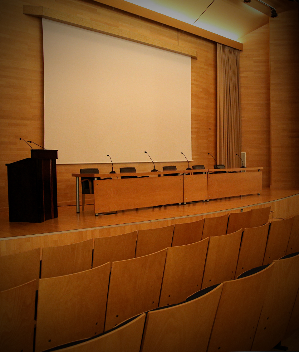 Salón de Actos con butacas a ambos lados y escenario con una mesa en donde hay micrófonos